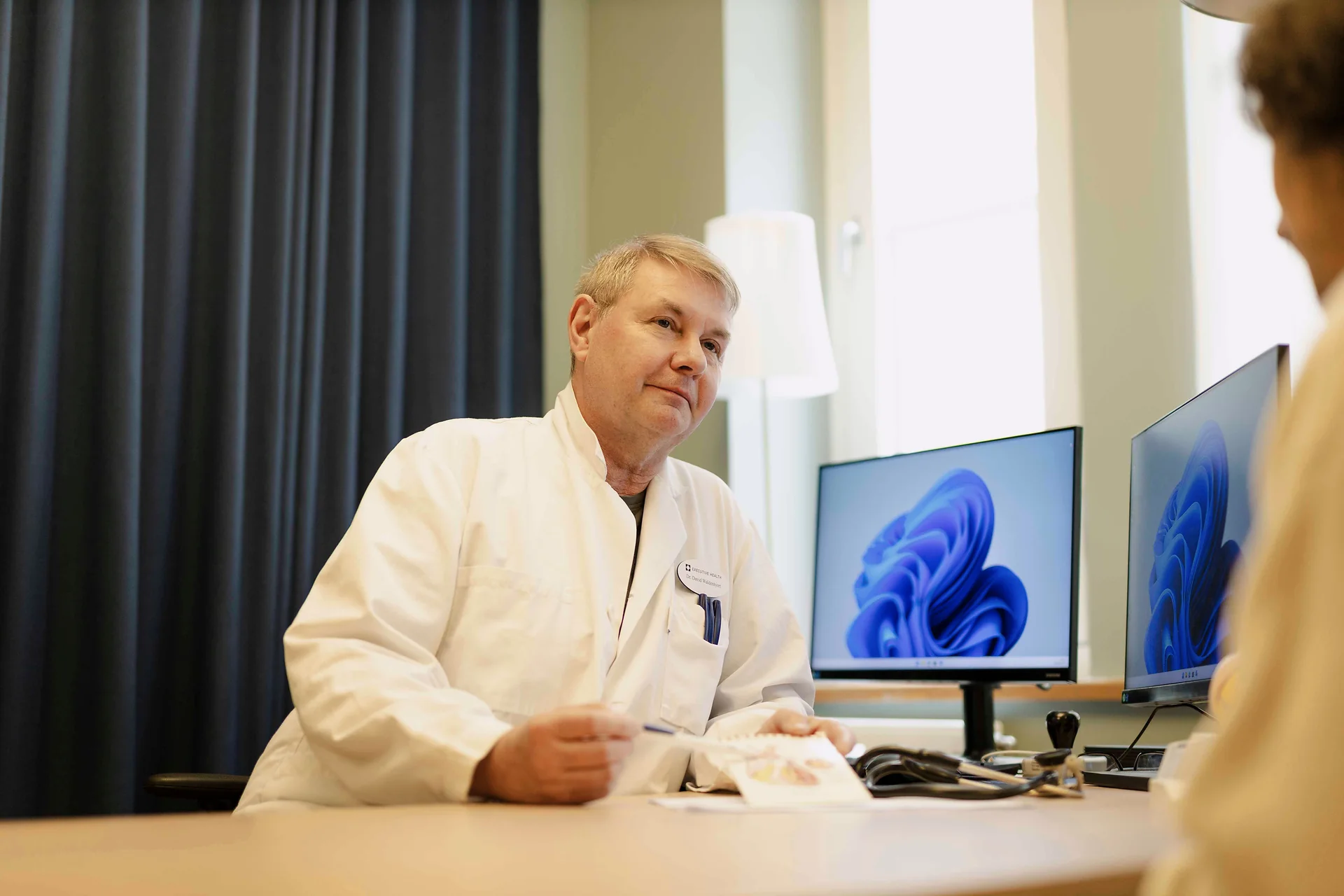 Executive Health doctor consulting with a patient in a clinical room with medical monitors.